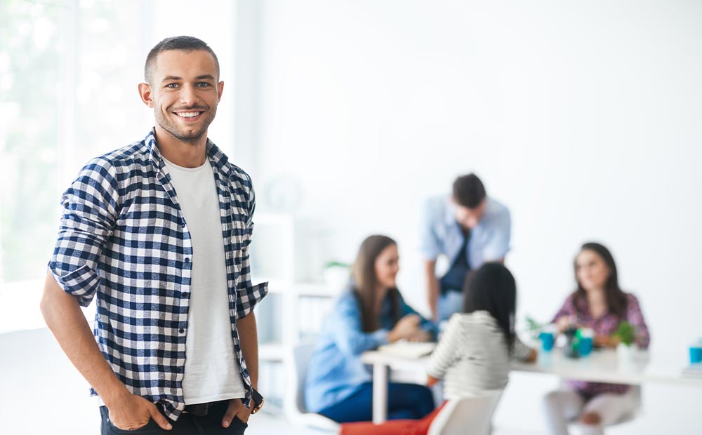 Joven empresario sonriendo a la cámara con un equipo reunido en el fondo.