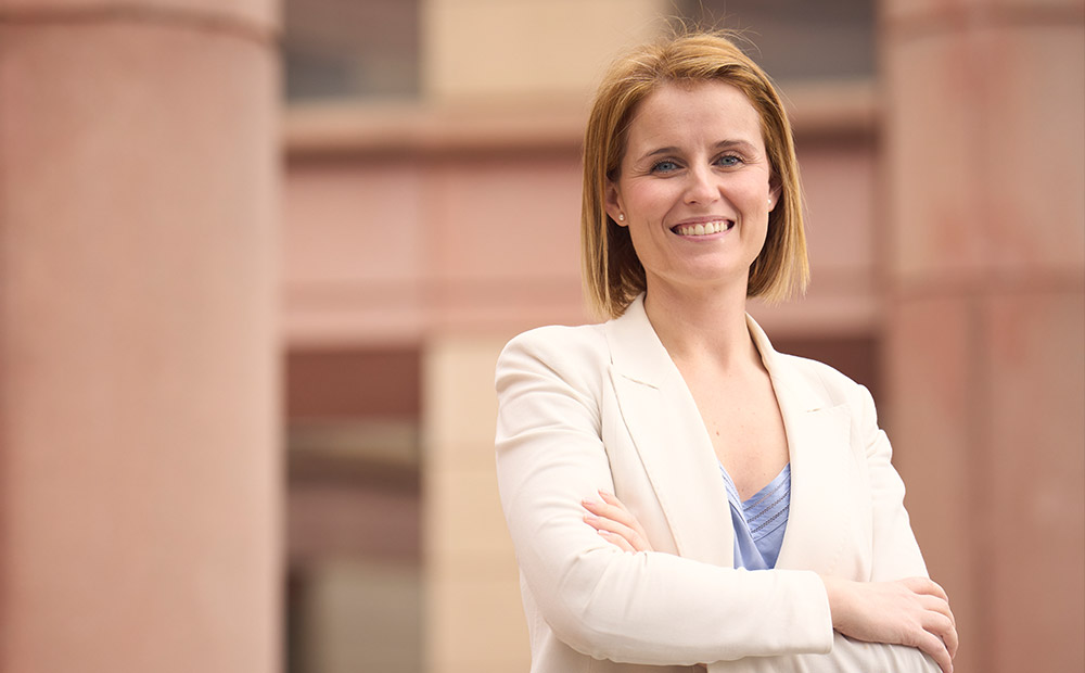 Mujer de pie con los brazos cruzados, vestida con un blazer blanco y una blusa azul, frente a una estructura arquitectónica imponente con grandes columnas. Su postura firme y profesional transmite seguridad y determinación, cualidades clave del empoderamiento femenino. Esta representación visual refuerza la idea de que las mujeres ocupan espacios de poder y autoridad, proyectando liderazgo y confianza en cualquier entorno, ya sea institucional, empresarial o social.