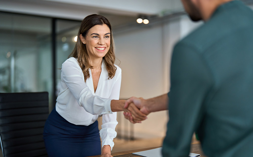 Dos personas se dan la mano en una oficina. Una está de pie y la otra sentada, en un entorno con pared de cristal y mobiliario de oficina. La escena representa un acuerdo o resolución en el ámbito del Derecho Laboral.