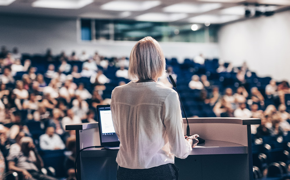 La imagen muestra a una mujer de pie frente a un atril, presentando a una audiencia en lo que parece ser una sala de conferencias o aula universitaria. Con un portátil sobre el atril y el público atento, esta escena representa la importancia de las mujeres en espacios de formación, liderazgo y visibilidad profesional.