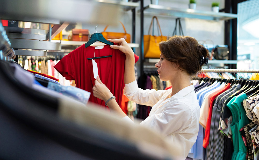 Una mujer en una tienda de ropa sosteniendo un vestido rojo en una percha, observándolo detenidamente, posiblemente revisando la etiqueta de precio. Esta escena ilustra el momento en que el consumidor evalúa el valor percibido de un producto, lo cual está directamente influenciado por la estrategia de fijación de precios utilizada por la marca. Factores como el posicionamiento, los costos, la competencia y la psicología del consumidor juegan un papel clave en cómo se determina ese precio final.
