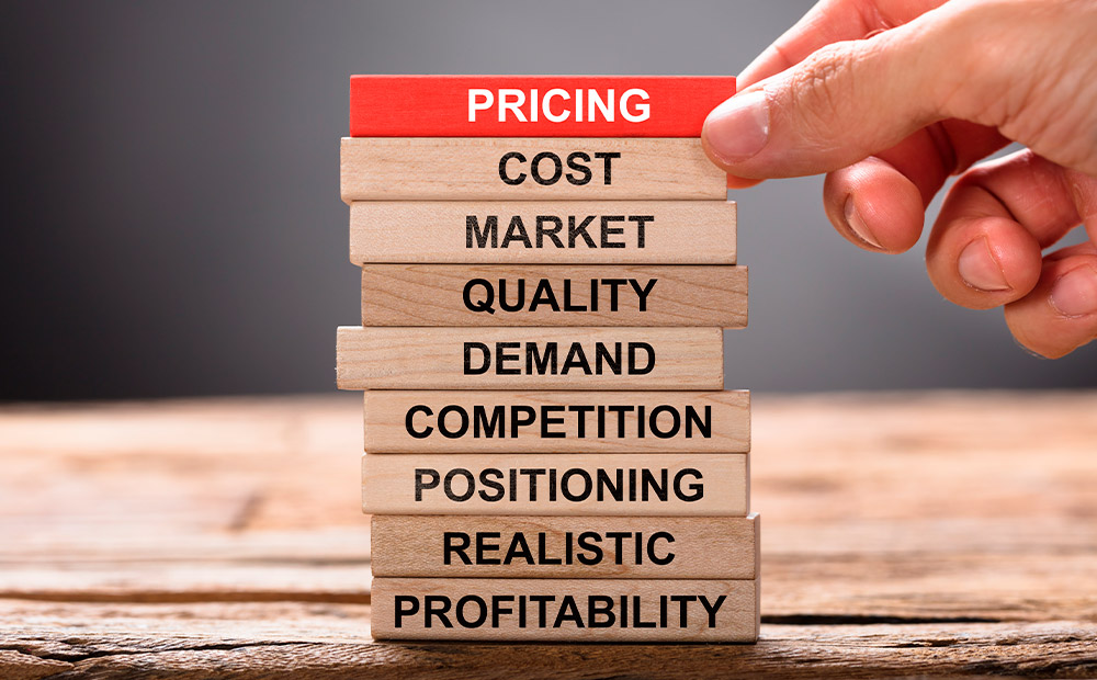 La imagen muestra una torre de bloques de madera apilados verticalmente, cada uno con una palabra clave relacionada con la estrategia de precios, como “COST”, “MARKET”, “QUALITY”, “DEMAND”, entre otros. En la parte superior, una mano coloca el bloque rojo con la palabra “PRICING”. Esta representación visual simboliza cómo la fijación de precios se construye sobre múltiples factores estratégicos, desde los costos y la competencia hasta la percepción de valor y la rentabilidad, destacando la importancia de un enfoque integral y bien fundamentado.
