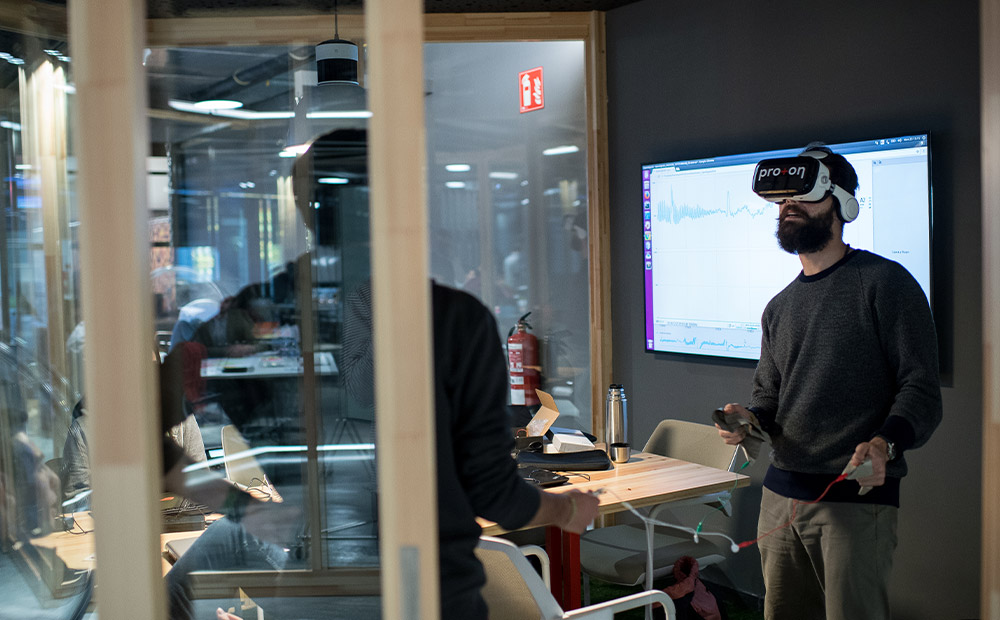 Se muestra una persona utilizando un visor de realidad virtual y mandos en una sala de reuniones, mientras en el fondo se proyectan gráficos en una pantalla grande. Este entorno tecnológico refleja cómo herramientas avanzadas como la realidad virtual, impulsadas por inteligencia artificial, están transformando la educación superior. La escena ilustra el potencial de estas tecnologías para mejorar la visualización de datos, fomentar el aprendizaje inmersivo y ofrecer experiencias educativas más efectivas e innovadoras.