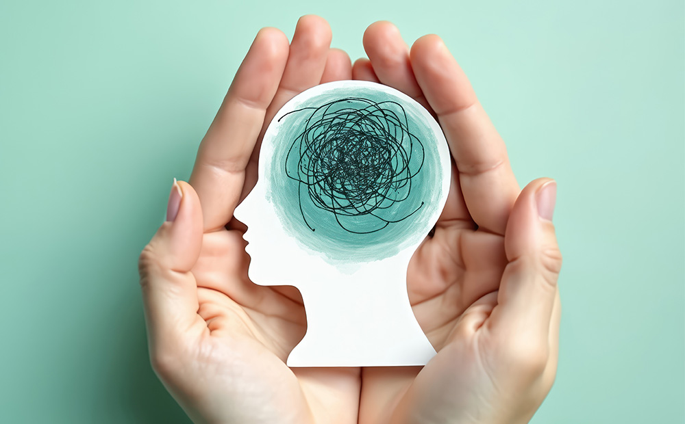 Dos manos sostienen con delicadeza la silueta de una cabeza humana que contiene un entramado caótico, simbolizando la confusión mental y el estrés. Esta imagen representa visualmente los procesos cognitivos alterados y destaca la importancia del cuidado de la salud mental en contextos personales y profesionales.