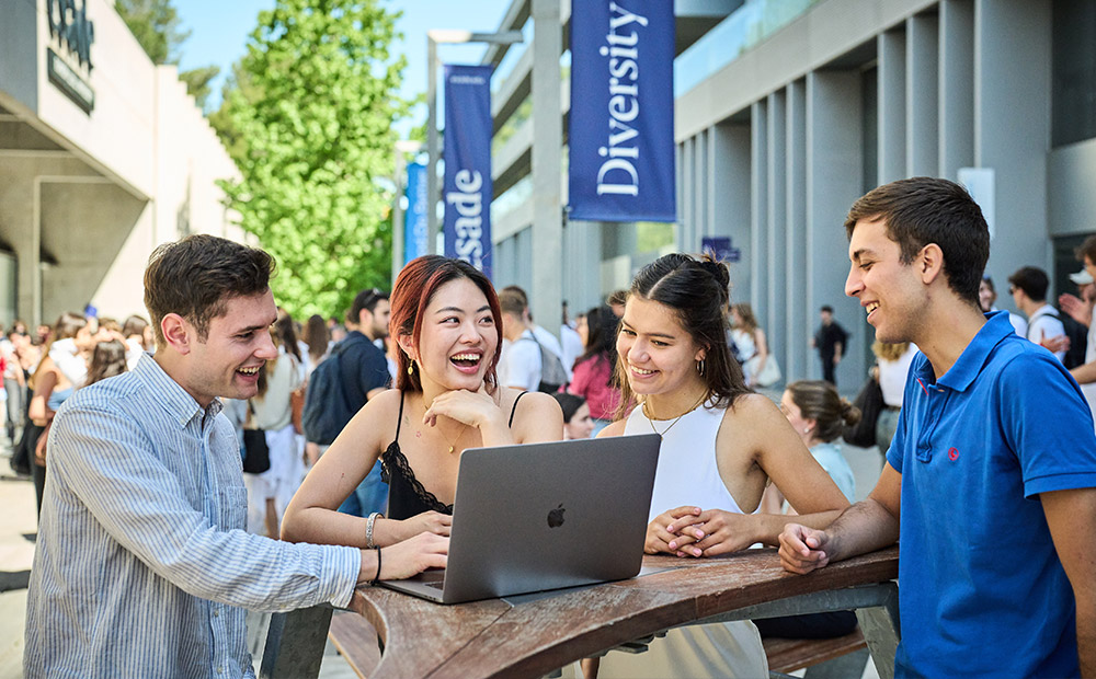 Vemos cuatro personas reunidas en campus al aire libre alrededor de un portátil, colaborando en un proyecto. Están en una mesa de madera, y en el fondo hay banderas con palabras como "Diversity" y "Engage".