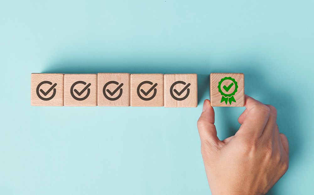 En la imagen se ven varios bloques de madera alineados, cada uno con un icono de verificación, mientras una mano coloca al final un bloque adicional con un distintivo verde que simboliza logro o certificación. 