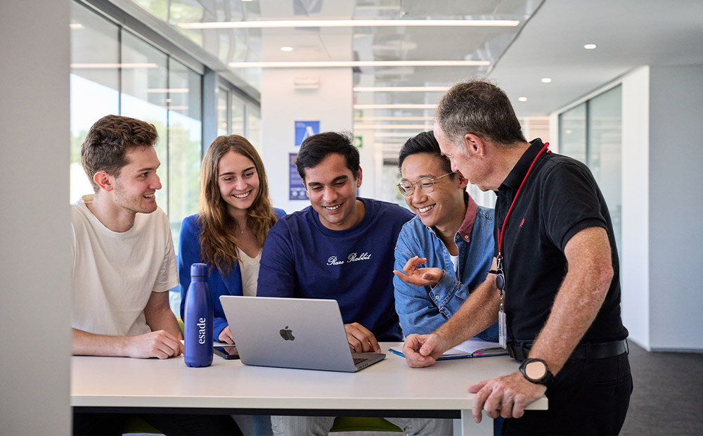 La imagen muestra a un grupo de estudiantes reunidos alrededor de una mesa en un campus universitario, trabajando con un ordenador portátil y conversando de manera colaborativa.