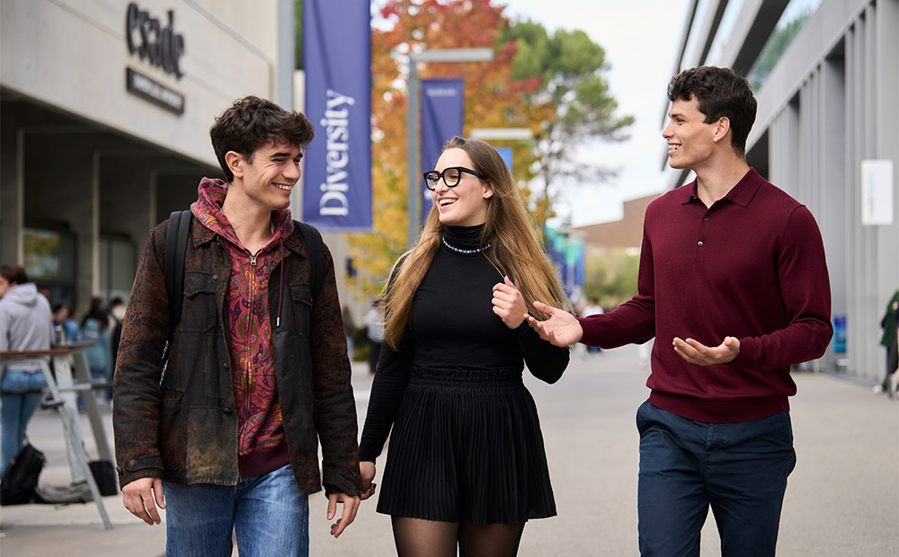 La imagen muestra a tres personas caminando por un campus universitario moderno, con edificios acristalados y banderolas visibles que incluyen la palabra “Diversity”.