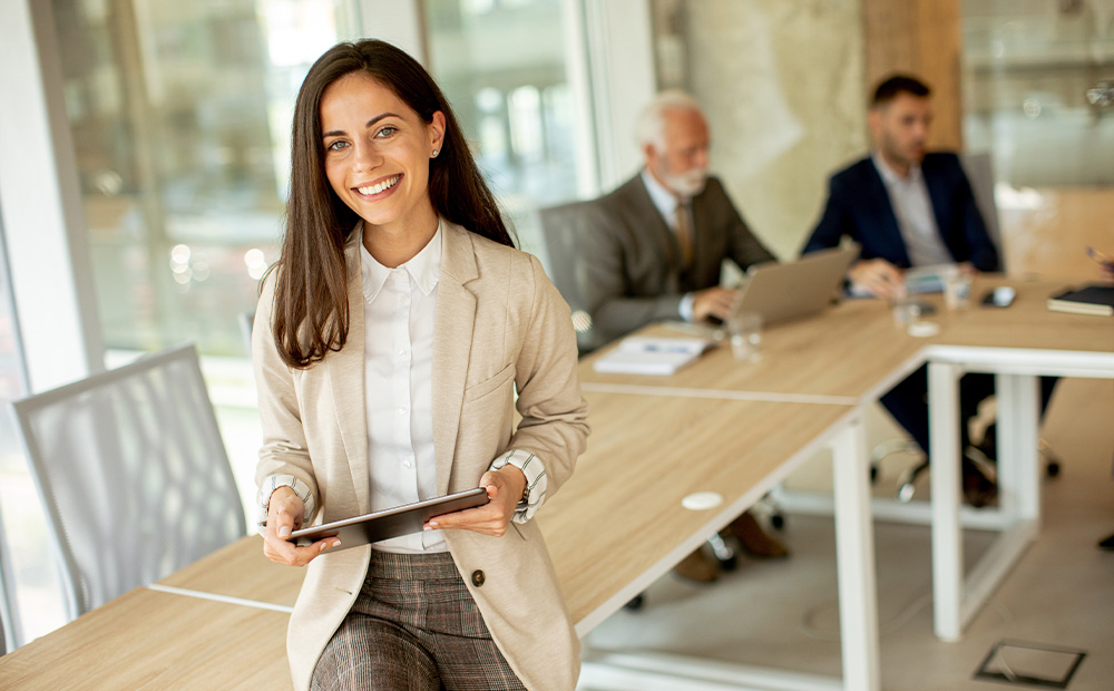 En la imagen se muestra a una profesional en una sala de reuniones moderna, sosteniendo una tableta digital mientras sus compañeros trabajan al fondo, transmitiendo un entorno corporativo en plena evolución.