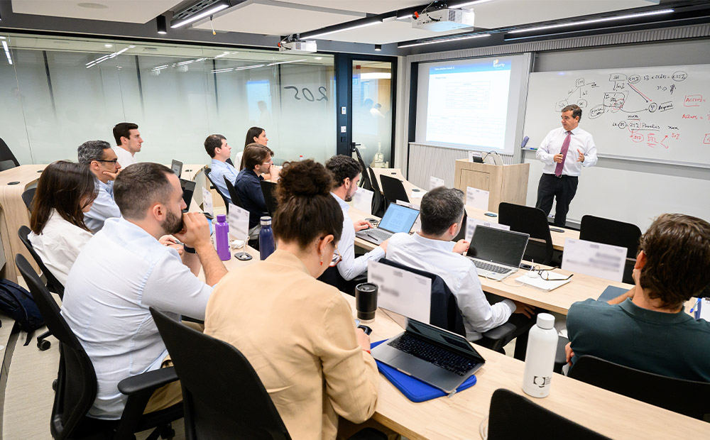 La imagen muestra una sesión de formación corporativa en un aula ejecutiva, donde un docente comparte conocimientos mientras los participantes trabajan con ordenadores y materiales de aprendizaje.