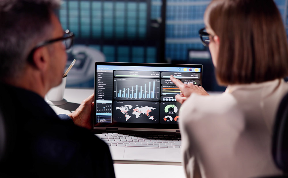 En una oficina, dos profesionales analizan conjuntamente un reporte en pantalla que incluye gráficos y mapas.