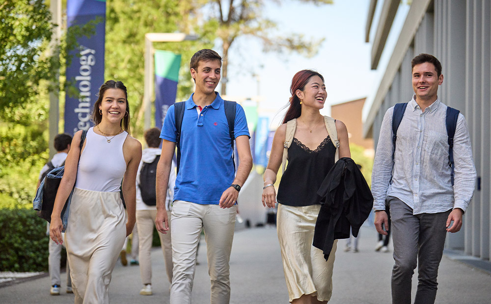 La imagen muestra a cuatro personas caminando al aire libre por un camino, con mochilas a la espalda y un entorno que sugiere un campus universitario, donde se observan edificios, árboles y pancartas con la palabra “technology”. Esta escena refleja la vida cotidiana de los estudiantes internacionales en España, quienes gracias a la Visa de Estudiante pueden integrarse en entornos académicos modernos, participar en actividades universitarias y disfrutar de una experiencia educativa completa dentro y fuera del aula.