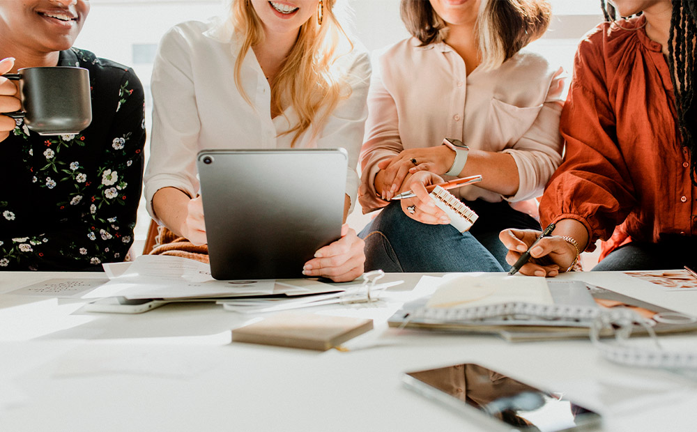 The image shows four women gathered around a table, actively collaborating in a professional setting. One holds a tablet, another a cup, and the table is scattered with notebooks, papers, and a mobile phone—elements that reflect a dynamic work environment. This scene illustrates spaces where women share ideas, lead projects, and develop their potential.