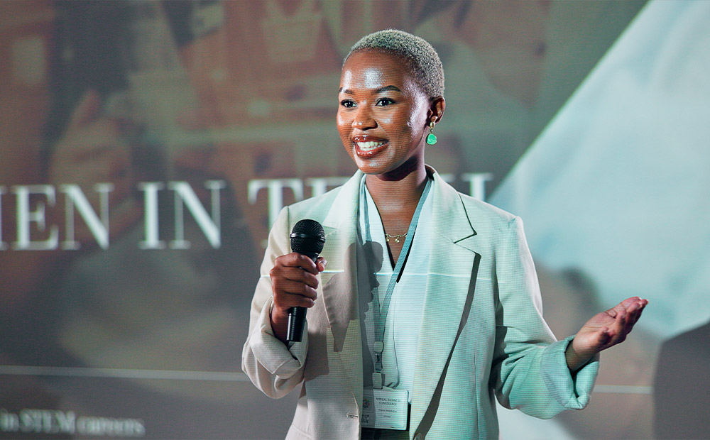 The image shows a woman standing at a podium, presenting to an audience in what appears to be a conference room or lecture hall. With a laptop on the podium and the audience engaged, this scene highlights the importance of women in spaces of education, leadership, and professional visibility.