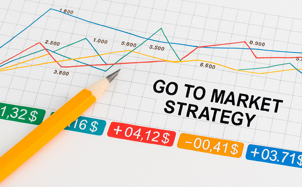 Un gráfico con líneas de colores que representan datos financieros a lo largo del tiempo, acompañado de cifras en euros y dólares, y el texto central "GO TO MARKET STRATEGY", junto a un lápiz amarillo que sugiere planificación activa.