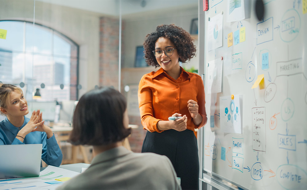 An executive presenting in front of a group in a modern office, using a whiteboard filled with notes, diagrams, and charts. This scene reflects essential skills for a marketing director, including leadership, strategic communication, and team management. 