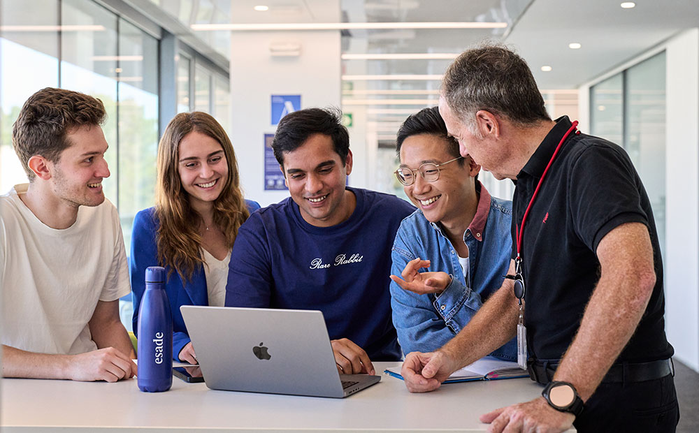 Five people are gathered around a laptop—students and a professor—in a modern, well-lit space, collaborating on an academic activity. This scene reflects the key role of artificial intelligence in fostering collaborative and personalized learning in higher education. The interaction between students and educators in digital environments shows how AI can improve educational quality, facilitate access to knowledge, and transform the university learning experience.