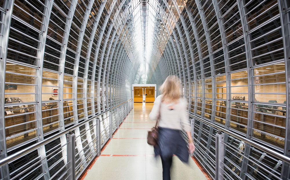 La imagen muestra la entrada de uno de los campus de Esade, es un pasillo con paredes de metal y vidrio, donde una persona camina hacia una puerta al fondo. Este entorno académico de diseño avanzado simboliza la transformación digital en la educación superior, destacando cómo la inteligencia artificial puede integrarse en espacios de aprendizaje para mejorar la eficiencia.