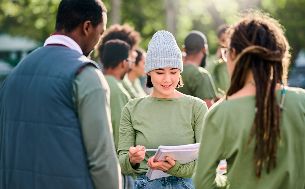 A group of people is gathered outdoors, with trees in the background, participating in a collaborative activity. In the foreground, three individuals are interacting, one of them holding a notebook, suggesting organization and planning. This scene reflects the fieldwork of many NGOs, where measuring social impact begins with direct observation, community engagement, and the collection of qualitative data to assess the real reach of their initiatives.