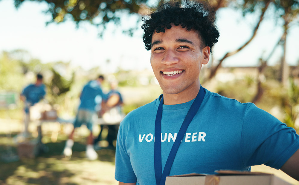 A person wearing a blue shirt with the word "VOLUNTEER" is holding a cardboard box and surrounded by others participating in a community activity outdoors. This scene represents volunteering as one of the most visible and valuable actions within an NGO, emphasizing the importance of measuring social impact not only through tangible results but also through civic engagement, participation, and the strengthening of social bonds.