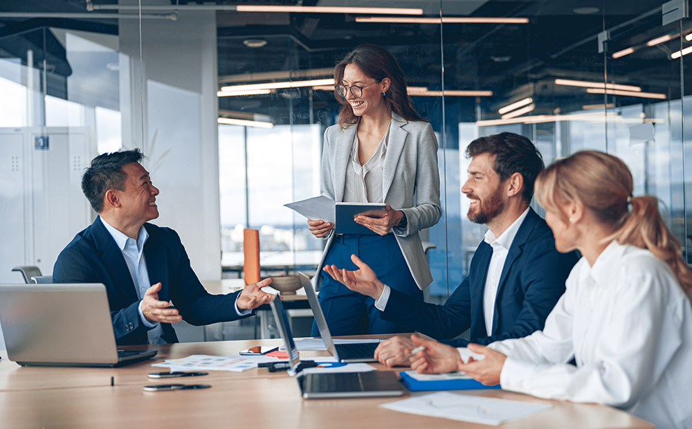 A group of five professionals gathered in a modern office around a conference table. They are engaged in collaborative work, surrounded by laptops, documents, and notepads. The space features large windows that let in natural light, enhancing the corporate and professional atmosphere.