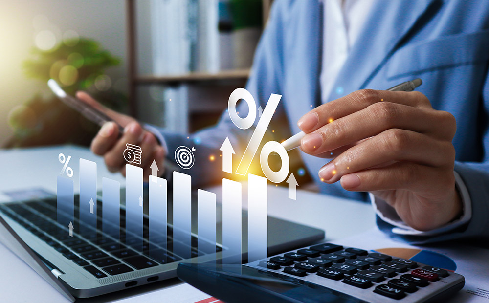 Una persona trabajando con un portátil, un móvil y una calculadora, mientras se proyectan digitalmente gráficos de barras y símbolos financieros que indican crecimiento y análisis.