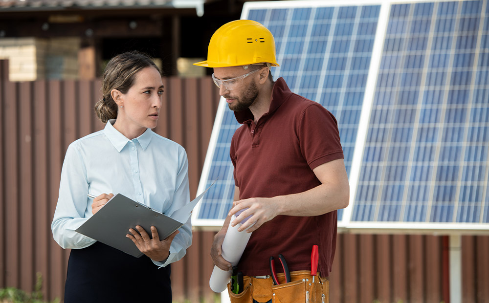 The image shows two professionals reviewing plans in front of large solar panels, symbolizing the practical application of sustainability principles in the energy sector.