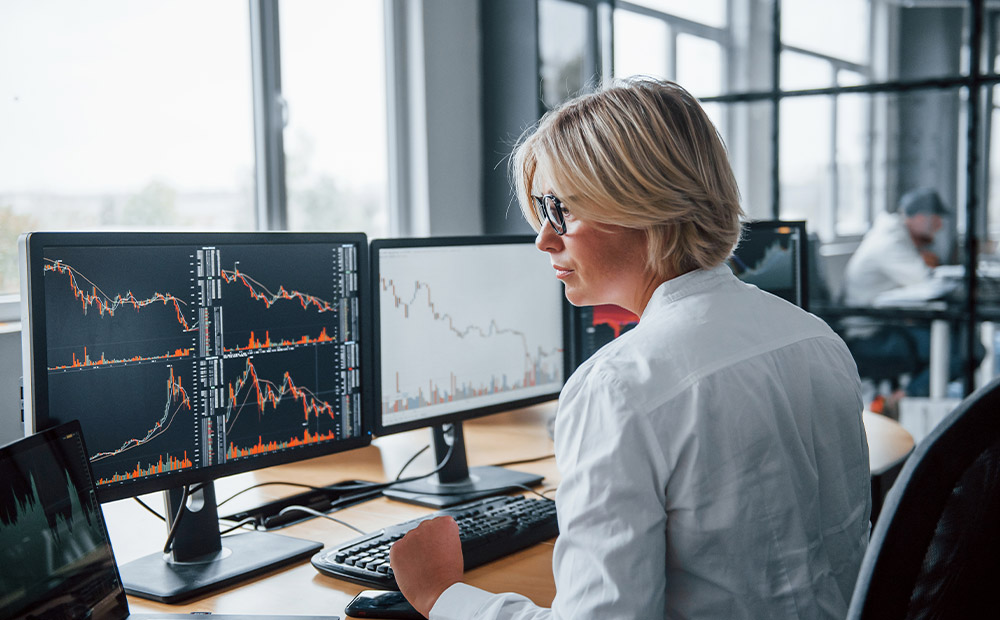The image shows an individual working in an office in front of multiple screens displaying financial charts with candlestick patterns and trading volumes, reflecting a professional environment focused on market analysis.
