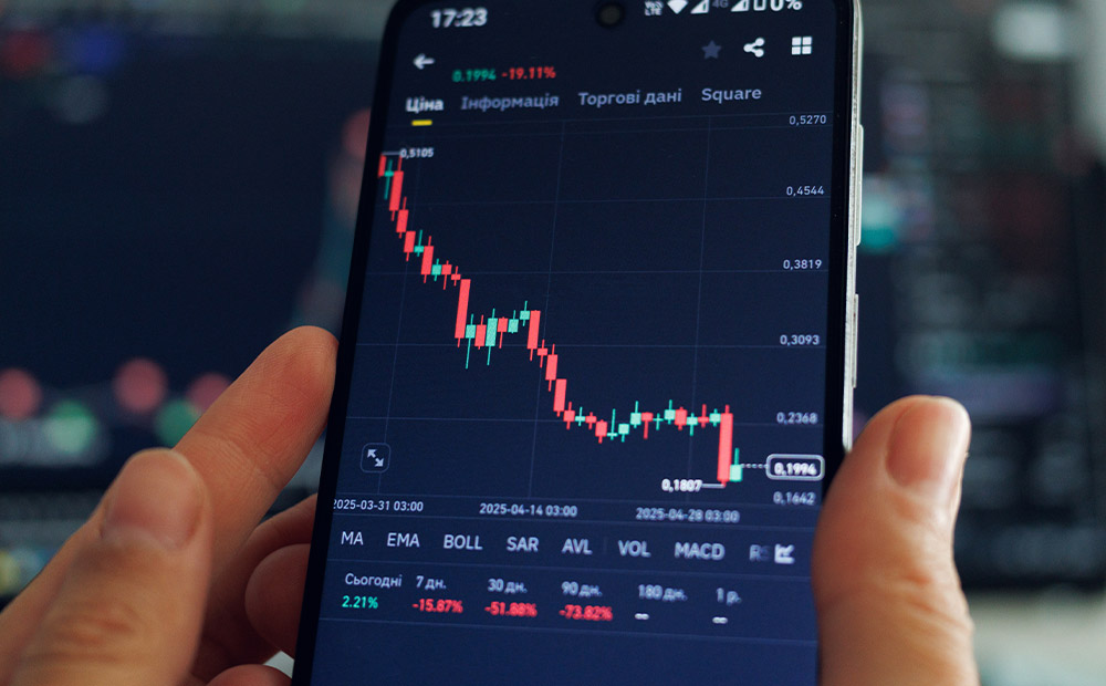 Vemos un teléfono móvil mostrando una plataforma de trading con un gráfico de velas japonesas en clara tendencia bajista, acompañado de indicadores técnicos como EMA, Bollinger Bands y MACD.