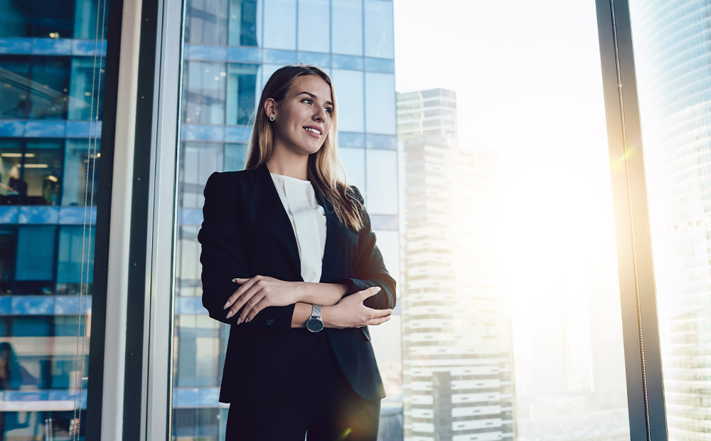 En la imagen se muestra a una profesional de pie en una oficina moderna, rodeada de grandes ventanales que dejan ver un skyline corporativo iluminado por la luz del sol, transmitiendo una sensación de crecimiento y proyección laboral.