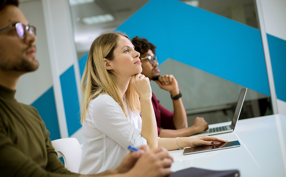 La imagen muestra a un grupo de personas en un aula moderna, tomando apuntes y utilizando un ordenador portátil, lo que refleja un entorno formativo dinámico y orientado al desarrollo profesional.