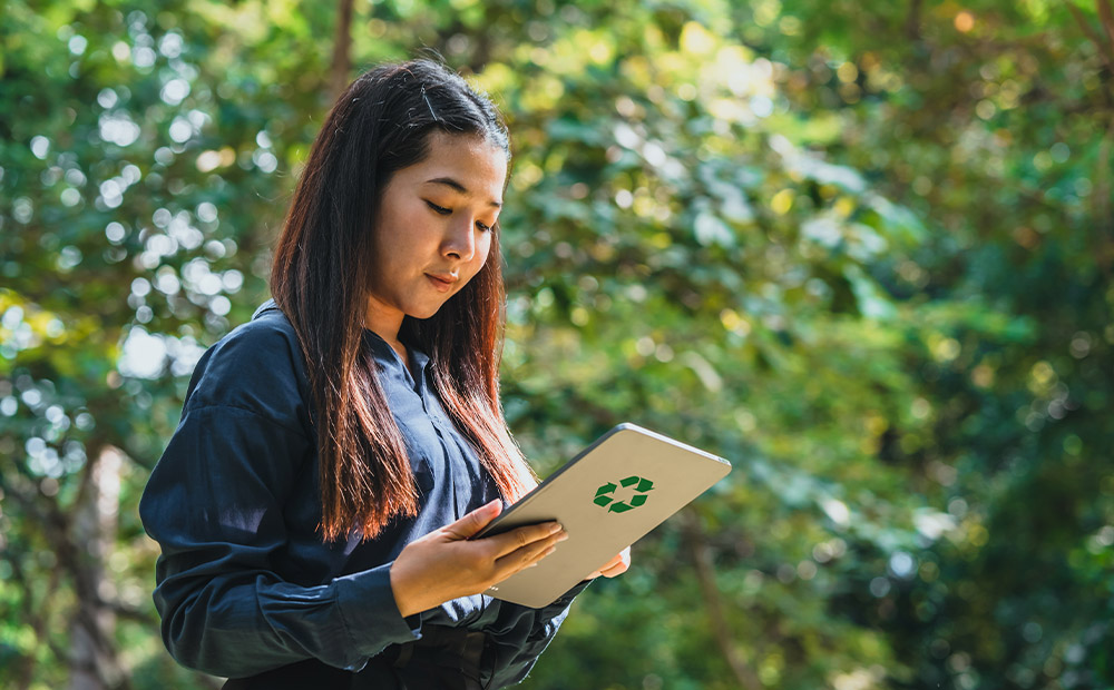 The image depicts a person outdoors, surrounded by abundant greenery, holding a tablet that displays the recycling symbol.