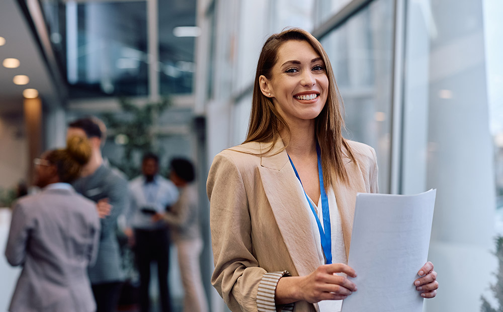 The image shows a person in a modern corporate setting holding several documents, while other professionals can be seen in the background engaging in conversations and networking activities.
