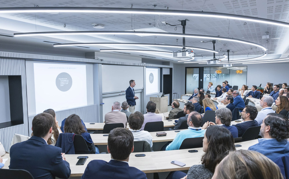 En la imagen se observa una sesión formativa para ejecutivos en una sala tipo auditorio moderno, donde un profesor experto expone contenidos estratégicos ante un público profesional atento, apoyado por una presentación visual.
